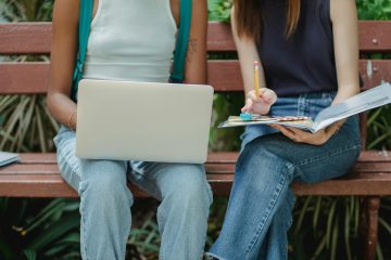 Two college students, young women, do homework together on a bench. One uses a laptop and the other uses a notebook.
